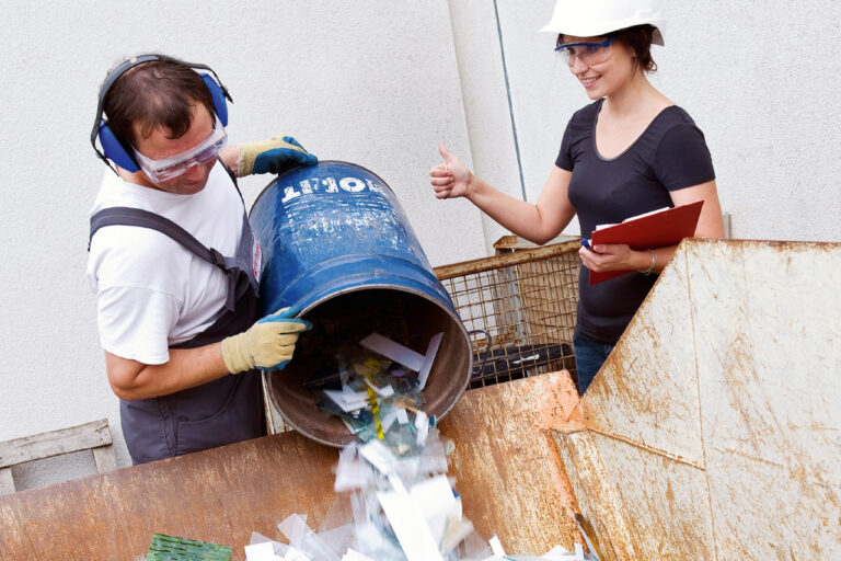 Ein Mann, der Handschuhe, Gehörschutz und Schutzbrille trägt, schüttet Glasbruch aus einer Tonne in einen Container. Eine Frau, die für Arbeitsschutz zuständig ist, gibt ihm ein Zeichen, dass er alles richtig macht.