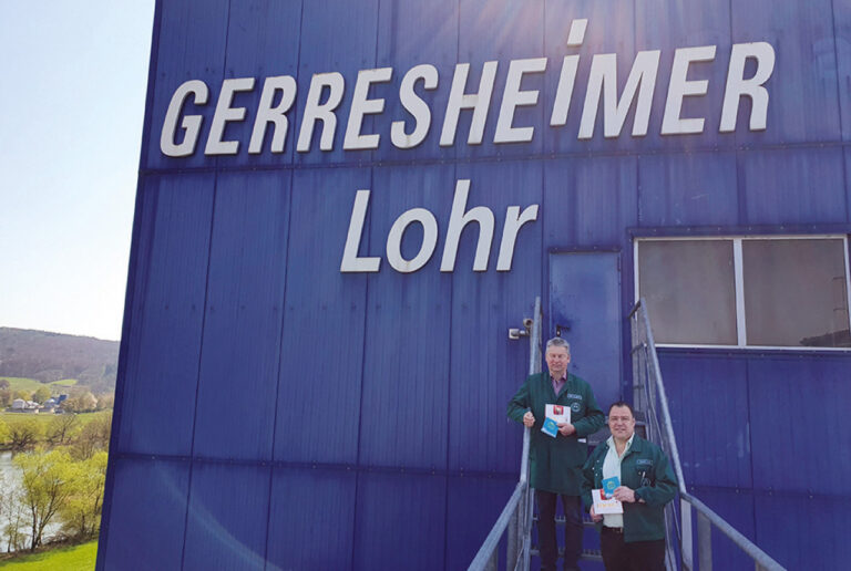 Die beiden, in der Bildunterschrift genannten Herren, stehen auf einer Außentreppe vor einer vollkommen blau gestrichenen Werkshalle, auf der das große weiße Firmenlogo angebracht ist.