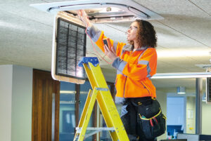 Ein Frau mit Schutzkleidung, Schutzbrille und Werkzeug steht auf einer Leiter in einem Großraumbüro. Sie arbeitet an einer Entlüftungsanlage.
