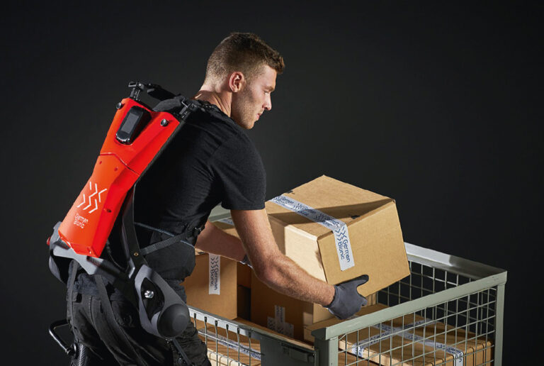 Ein Mann, der ein schwarz-rotes Exoskelett trägt, hebt schwere Kartons aus einer Gitterbox.