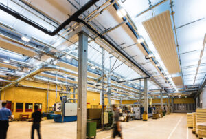 Eine große Fabrikhalle mit etlichen Röhren, Leitungen und vielen hellen Deckenleuchten in großer Höhe entlang des Dachs der Halle. Einige Mitarbeiter bewegen sich beschäftigt in der Halle.