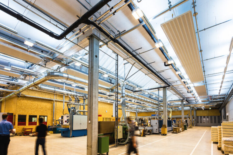 Eine große Fabrikhalle mit etlichen Röhren, Leitungen und vielen hellen Deckenleuchten in großer Höhe entlang des Dachs der Halle. Einige Mitarbeiter bewegen sich beschäftigt in der Halle.