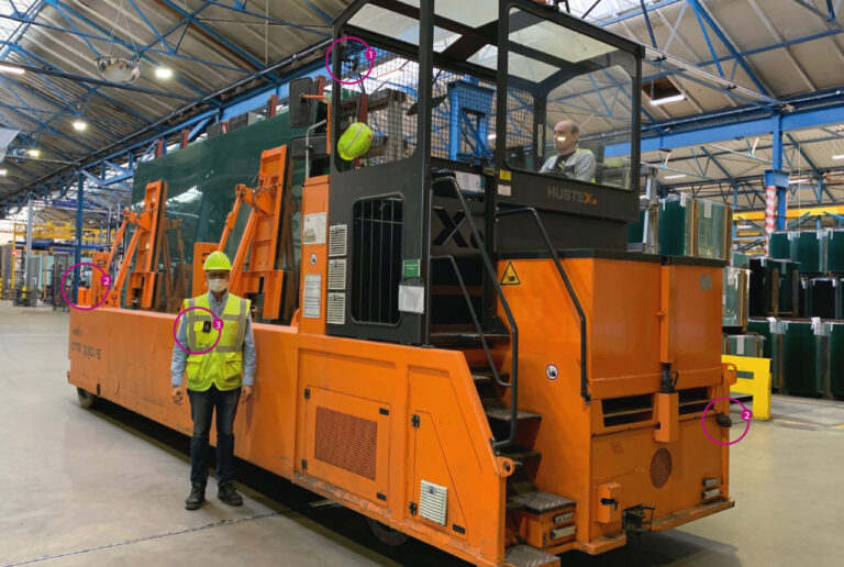 Blick in eine Lagerhalle: Mann mit gelber Weste, gelbem Schutzhelm und Mundschutz steht mittig vor einen sehr großen Transportmaschine für Glas. Ein Fahrer sitzt im Führerhaus der Maschine oberhalb der Kopfhöhe des anderen Manns. Er kann ihn nicht sehen. Mit zwei lila Kreisen sind Empfänger und ein Display für den Fahrer an beiden Enden der Maschine markiert. Ein dritter Kreis markiert einen Sender, den der Mann vor der Maschine in Brusthöhe an der Weste trägt. Die Kreise markieren auf dem Bild die Funktionsweise eines Personenerkennungssystems: Der Fahrer wird damit vor Personen außerhalb seines Blickfeldes automatisch gewarnt.