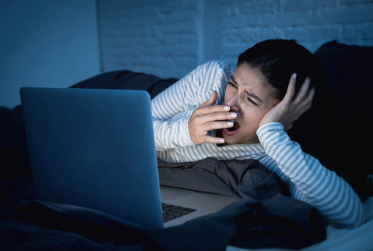 eine junge Frau liegt abends oder nachts im Bett vor einem erleuchteten Laptop Bildschirm und gähnt intensiv mit der Hand vor dem M