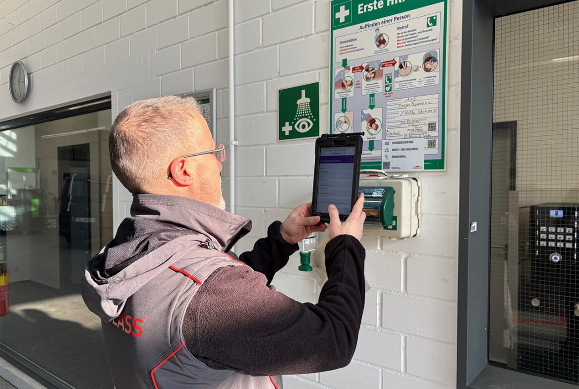 Ein Mitarbeiter scannt mit einem Tablett den QR-Code am Erste-Hilfe-Bord.