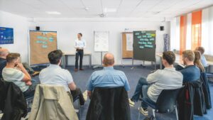 Ein Führungskräftetraining mit Vortrag vor Flipcharts. Ein Kreis von Teilnehmern hört zu.